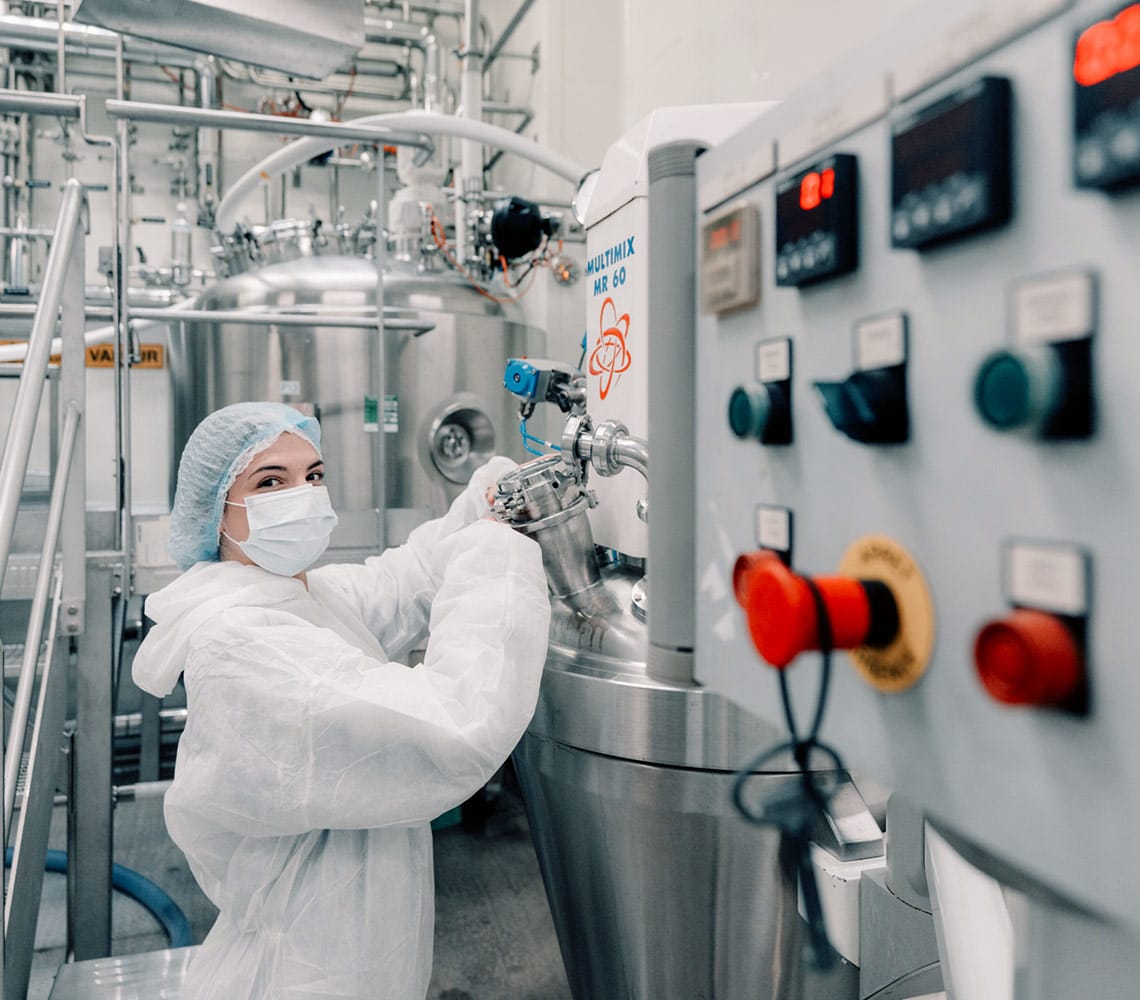A woman in a white coat and surgical mask operating machinery in a laboratory.